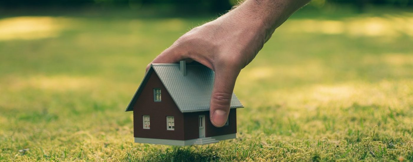 Concept of selling a house. A hand is holding a model house above green meadow.