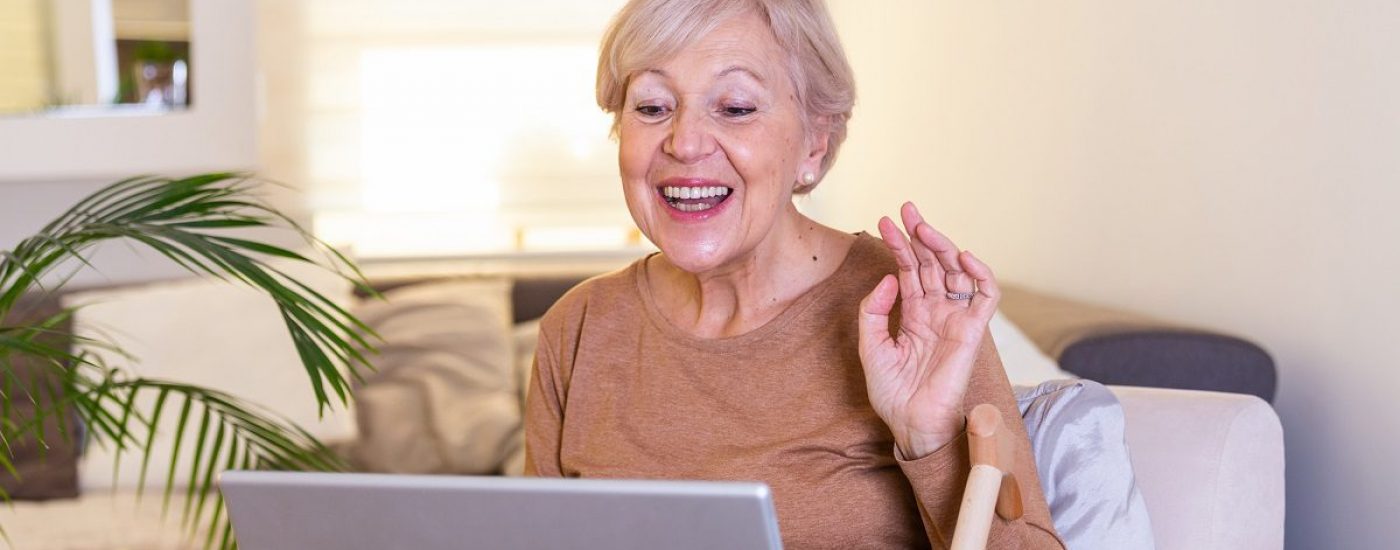 Happy mature woman waving to someone while having a video call over laptop at home. Gray-haired senior woman waving hand in front of laptop while having video call with her family members.