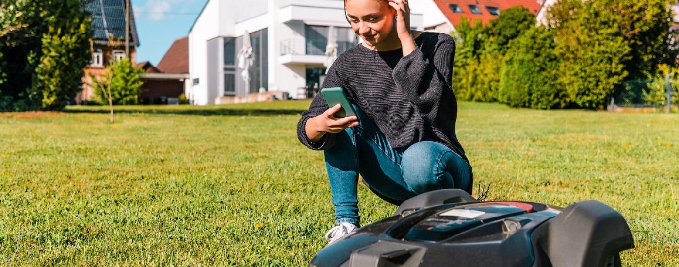 Young woman controlling a robot lawn mower with her smart phone in the summer. The woman kneeling next to the robotic mower, looking to her smart phone to program the robot mower. Modern Home in the background.