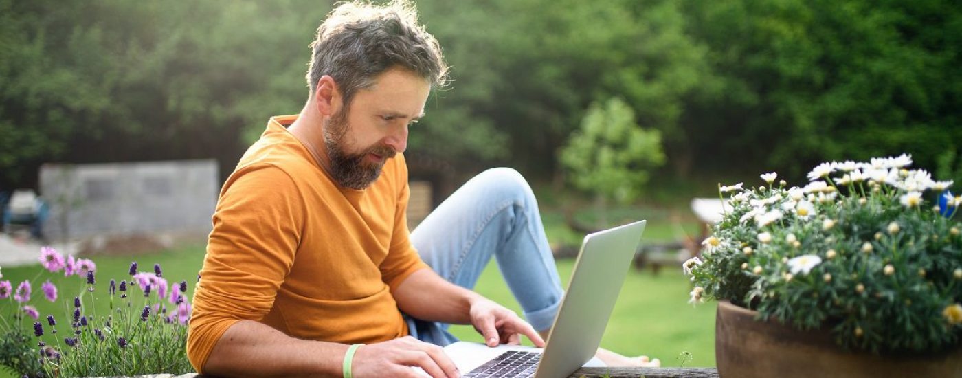 Happy mature man with laptop working outdoors in garden, home office concept.