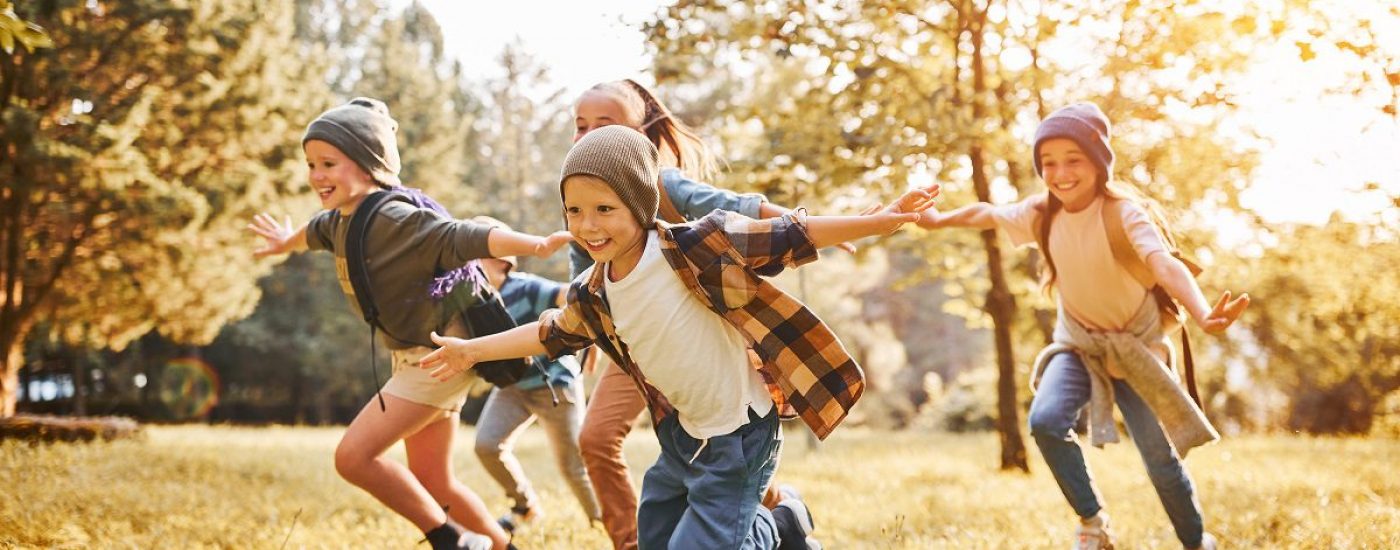 Group of happy joyful school kids with backpacks running with outstretched arms in forest on sunny spring day, excited children scouts boys and girls having fun during camping activity in nature